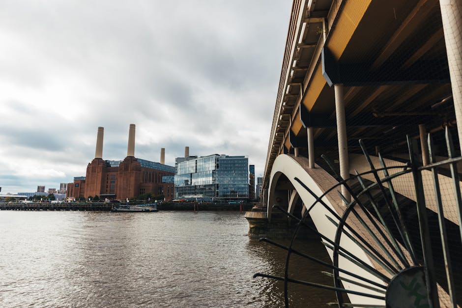 A large industrial building, resembling the former Battersea Power Station, constructed of red brick with distinctive tall white chimneys rising above it, situated near a river. The foreground feature