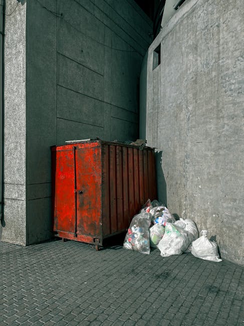 A red, weathered metal rubbish skip positioned against a dark green wall with a textured finish, adjacent to a light gray concrete wall. The skip has visible rust marks and a slightly dented surface,