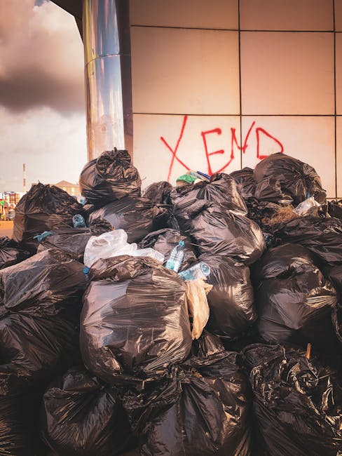 A red, weathered metal rubbish skip positioned against a dark green wall with a textured finish, adjacent to a light gray concrete wall. The skip has visible rust marks and a slightly dented surface,
